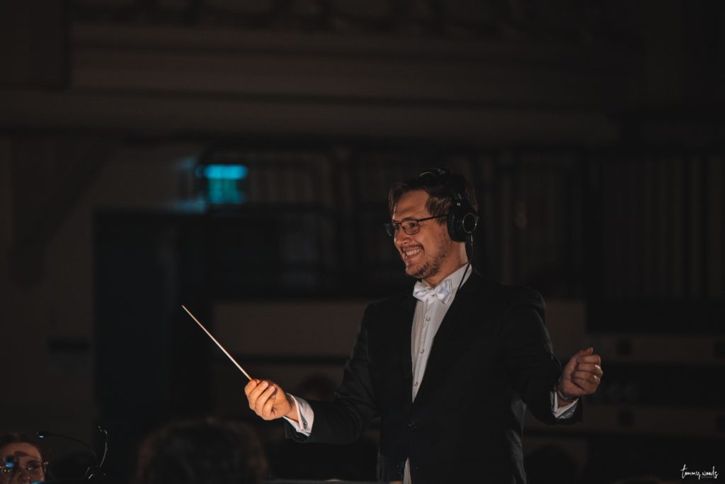 Artistic Director Dr Nathan Cummins holding his baton in an outstretched arm while smiling.